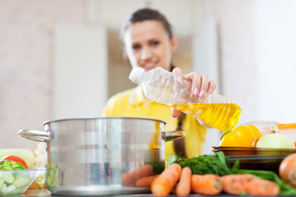 Woman in yellow pours oil from bottle into the pan. Focus on bottle